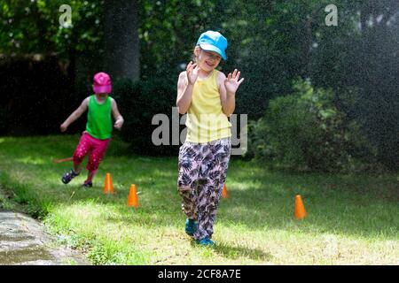 Kleine Kinder und Sommer Outdoor-Aktivitäten für Familien. Zwei lachende Schwestern, glückliche Grundschüler, die draußen spielen, von Wasser, Tropfen, Nebel besprüht Stockfoto