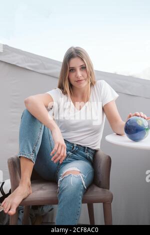 Lächelnde, lässige Frau in zerrissenen Jeans und weißem T-Shirt, die sich im Stuhl entspannt und in die Kamera schaut Stockfoto