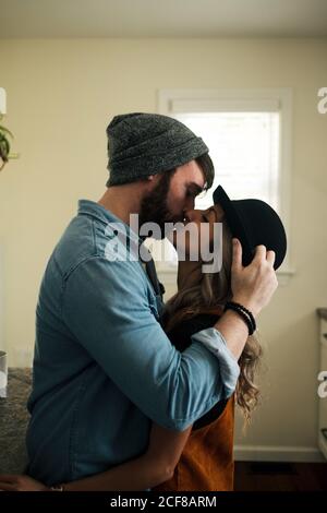 Seitenansicht einer fröhlichen Frau im Hut, die ihren Freund in einer Denim-Jacke küsst, während sie in der Küche mit geschlossenen Augen steht Stockfoto