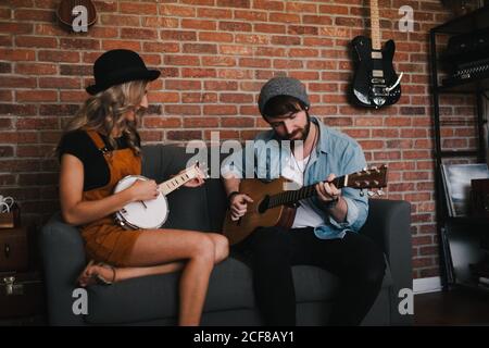 Lächelnde Frau, die mit gekreuzten Beinen auf dem Sofa sitzt und in Jeansjacke und Sockenmütze neben ihrem Freund Banjo spielt und am Wochenende akustische Gitarre spielt Stockfoto