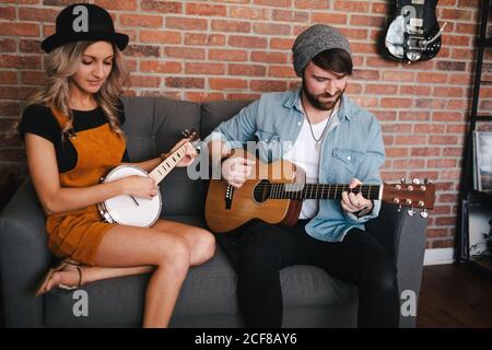 Lächelnde Frau, die mit gekreuzten Beinen auf dem Sofa sitzt und in Jeansjacke und Sockenmütze neben ihrem Freund Banjo spielt und am Wochenende akustische Gitarre spielt Stockfoto