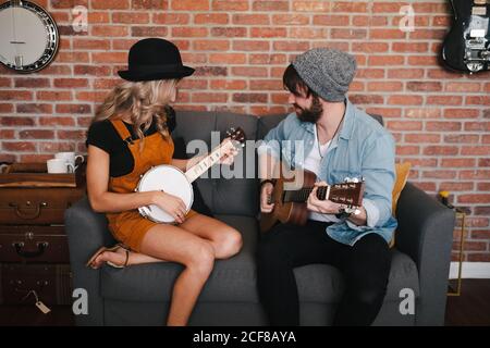 Lächelnde Frau, die mit gekreuzten Beinen auf dem Sofa sitzt und in Jeansjacke und Sockenmütze neben ihrem Freund Banjo spielt und am Wochenende akustische Gitarre spielt Stockfoto