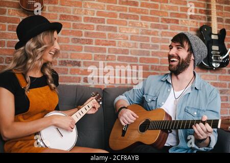 Lächelnde Frau, die mit gekreuzten Beinen auf dem Sofa sitzt und sich anschaut und am Wochenende in Denim-Jacke und Sockenmütze neben ihrem Freund Banjo spielt, um akustische Gitarre zu spielen Stockfoto