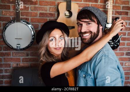 Seitenansicht von Boyfriend in Jeansjacke und Sockenmütze Kuscheln Freundin in schwarz Hut Blick auf Kamera auf gemütlich Couch in der Nähe von Ziegelwand mit Gitarren und Banjo in flach Stockfoto