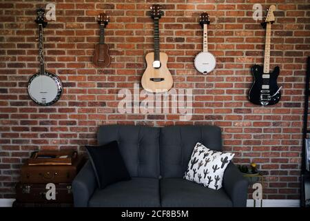 Sammlung von Gitarren und Banjos, die oben an der Ziegelwand hängen Gemütliches Sofa mit Kissen und Nachttisch beiseite in der Wohnung Stockfoto