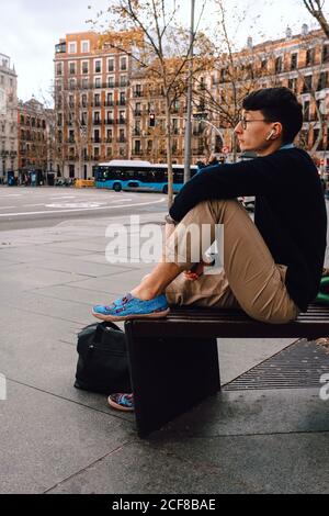 Ganzer Körper von asiatischen jungen trendigen männlichen in stilvollem Outfit Und Brillen sitzen auf kleinen Steinsäule posiert und schaut Entfernt in der Nähe eines alten, wunderschönen Gebäudes an der Straße Stockfoto