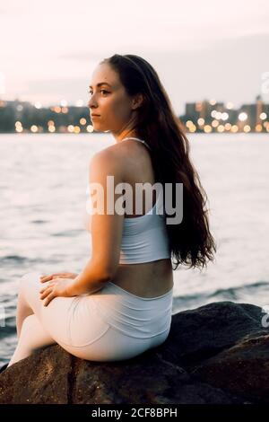 Seitenansicht des konzentrierten Weibchen in aktivem Ohr auf Stein sitzend Am Wasser beim Yoga vor dem Hintergrund der abendlichen Stadtlandschaft Und wegschauen Stockfoto