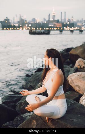 Von oben Seitenansicht der konzentrierten Weibchen im aktiven Sitzen Auf Stein am Wasser, während Yoga auf Hintergrund üben Abends Stadtbild und Blick weg Stockfoto