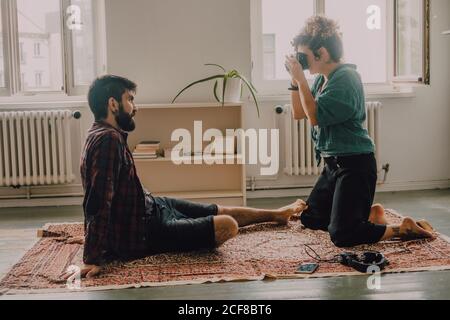 Seitenansicht einer Hipster-Frau, die einen Mann fotografiert, der barfuß sitzt und auf dem Wohnungsboden posiert Stockfoto