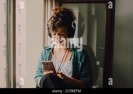 Lächelnde lockige Frau in Kopfhörern, die Musik hört, während sie auf dem Smartphone surft und auf dem Fensterbrett in der Wohnung sitzt Stockfoto