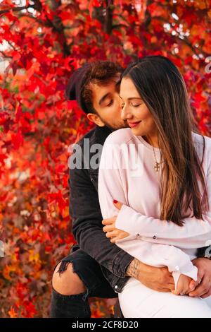 Glücklicher junger Mann und Frau umarmen und küssen, während sie auf der Bank in der Nähe des bunten Herbstbaums im Park sitzen Stockfoto