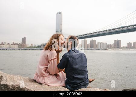 Seitenansicht des liebevollen Paares Umarmung und Küssen während des Sitzens Auf Steinen in der Nähe des Flusses auf dem Hintergrund der Brooklyn Brücke während Stadtbummel Stockfoto