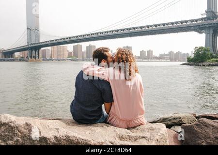 Rückansicht des liebevollen Paares Umarmung und Küssen während des Sitzens Auf Steinen in der Nähe des Flusses auf dem Hintergrund der Brooklyn Brücke während Stadtbummel Stockfoto