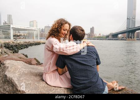 Seitenansicht des liebevollen Paares Umarmung und Küssen während des Sitzens Auf Steinen in der Nähe des Flusses auf dem Hintergrund der Brooklyn Brücke während Stadtbummel Stockfoto