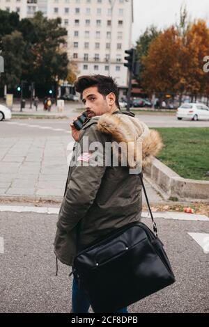 Seitenansicht des trendigen jungen Mannes, der auf dem Handy spricht Beim Wandern auf der Straße am Herbsttag Stockfoto