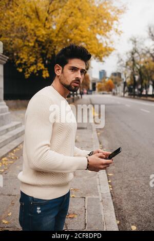 Seitenansicht des trendigen jungen Mannes, der mit Handy steht Am Straßenrand und im Herbst Auto fangen Stockfoto