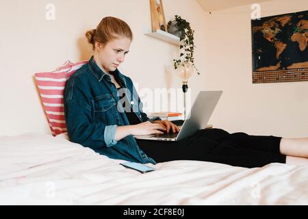 Fokussierte, entspannte junge Frau in legerer Kleidung, die auf einem weichen Bett sitzt, während sie den Laptop an einer hellbeigen Wand in einer gemütlichen, modernen Wohnung benutzt Stockfoto