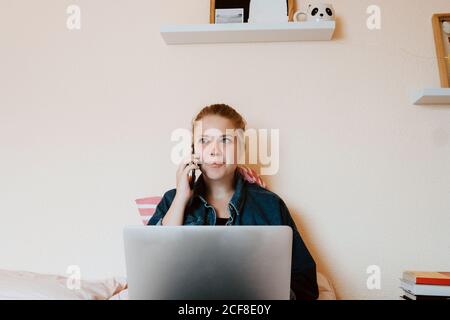 Positive entspannte junge Frau in lässiger Kleidung suchen weg, während Sitzen mit Laptop auf weichem Bett und Telefonieren In gemütlichem Schlafzimmer Stockfoto