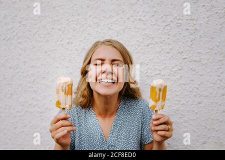 Frau mit geschlossenen Augen macht lustige Grimace Gesichter mit lecker Eislollies auf Stöcken auf weißem Hintergrund Stockfoto