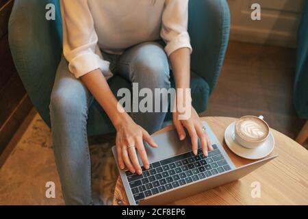 Von oben beschnitten unerkennbare junge Freiberuflerin in legerer Kleidung Bei einem leckeren heißen Getränk an einem runden Holztisch Und mit Netbook in hellen modernen Café Stockfoto