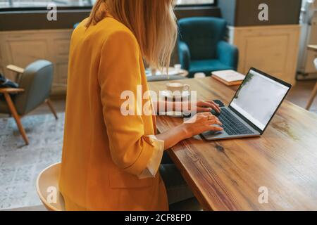 Cropped unkenntlich junge Freiberuflerin in legerer Kleidung mit lecker Heißes Getränk, während Sie am Holztisch sitzen und einen Laptop benutzen In hellem, modernem Café Stockfoto