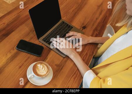 Von oben beschnitten unerkennbare junge Freiberuflerin in legerer Kleidung Mit einem leckeren heißen Getränk, während Sie an einem Holztisch sitzen und Mit Laptop in hellen modernen Café Stockfoto