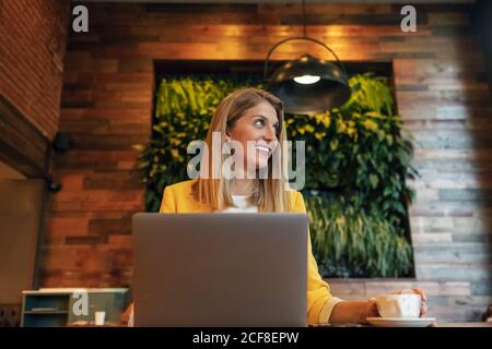 Erwachsene überfröhliche Geschäftsfrau in formeller Kleidung, die wegschaut und lächelt, während sie am Holztisch mit Laptop im modernen Café-Shop sitzt Stockfoto