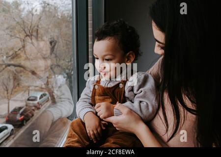Seitenansicht der liebevollen Mutter in legerer Kleidung hält niedlich Kleiner Sohn auf Händen, während er zu Hause neben dem Fenster stand Und wegzeigen Stockfoto