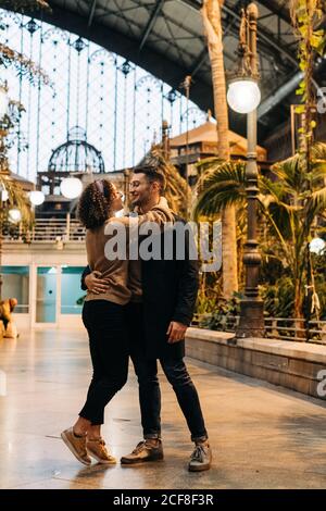 Fröhlicher junger Mann und Frau umarmen sich und schauen einander an, während sie während des Dates im beleuchteten Pavillon stehen Stockfoto