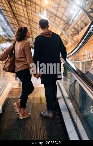 Rückansicht eines jungen Mannes und einer jungen Frau, die während des Dates in einem hell erleuchteten Einkaufszentrum die Hände halten, während sie auf einem beweglichen Gehweg stehen Stockfoto