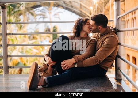 Fröhlicher junger Mann und Frau umarmen und küssen einander, während sie während des Dates im beleuchteten Pavillon sitzen Stockfoto