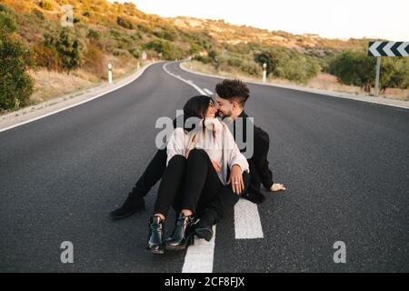 Hoher Winkel der modernen jungen ethnischen Paar in lässiger Kleidung Umarmung beim Sitzen auf leerer Asphaltstraße Stockfoto