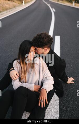 Hoher Winkel der modernen jungen ethnischen Paar in lässiger Kleidung Umarmung beim Sitzen auf leerer Asphaltstraße Stockfoto