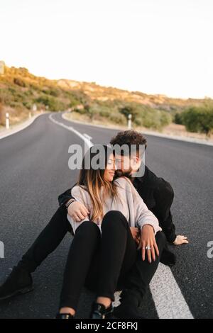 Hoher Winkel der modernen jungen ethnischen Paar in lässiger Kleidung Umarmung beim Sitzen auf leerer Asphaltstraße Stockfoto