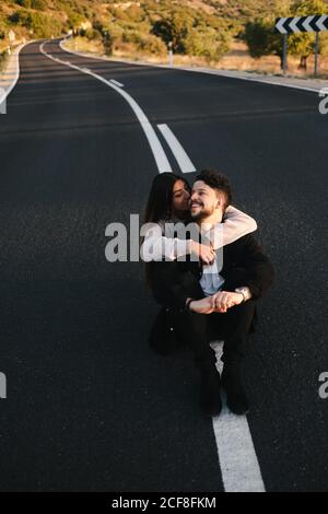 Hoher Winkel der modernen jungen ethnischen Paar in lässiger Kleidung Umarmung beim Sitzen auf leerer Asphaltstraße Stockfoto