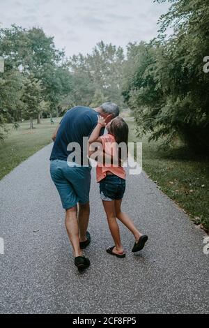 Rückansicht des präteen Mädchen, das Geheimnis erklärt und flüsternd innen Ohr des Vaters beim gemeinsamen Spaziergang auf dem Weg in grün park im Sommer Tag Stockfoto