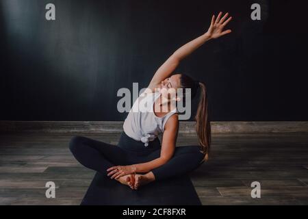 Junge entspannte Frau mit geschlossenen Augen und Arm angehoben sitzen In baddha konasana Position auf Sportmatte und Yoga üben In dunkler Halle Stockfoto