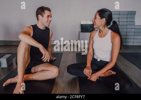 Junger Mann und eine Frau mit Barfußball in Sportkleidung lächeln, während sie sich gegenseitig anguckten und sich unterhielten, während sie auf Sportmatten auf dem Boden saßen und sich nach dem Yoga-Training im zeitgenössischen Studio ausruhten Stockfoto
