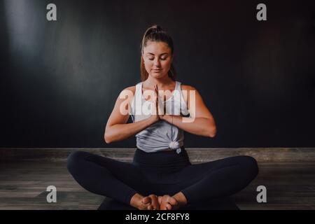 Ruhige, entspannte Frau mit geschlossenen Augen und namaste sitzend baddha konasana Position auf Sportmatte und Konzentration beim Üben Yoga im dunklen modernen Studio Stockfoto