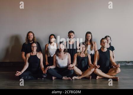 Der hohe Winkel der Gruppe der jungen ruhigen Frauen und der Männer In Sportbekleidung mit geschlossenen Augen und Gyan Mudra entspannend und Meditieren, während Sie in Padmasana-Position gegen weiße Wand sitzen Modernes Yoga-Studio Stockfoto