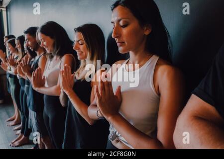 Gruppe von glücklichen jungen sportlichen Menschen in aktiven mit Augen Geschlossen entspannend und tiefes Atmen, während man mit Namaste im Stehen steht tadasana und meditieren neben schwarzer Wand im modernen Yoga-Studio Stockfoto