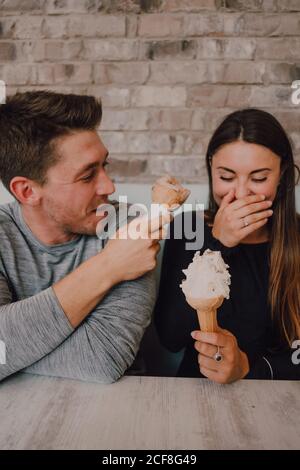 Ein fröhlicher Mann und eine junge Frau in legerer Kleidung, die sich mit leckerem Eis angucken, während sie am Tisch auf dem Sofa sitzen und sich in einem modernen Café im Loft-Stil entspannen Stockfoto