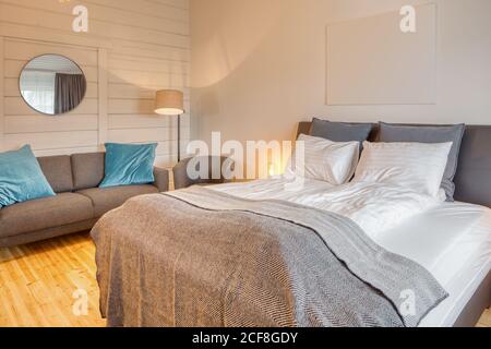 Komfortables Schlafzimmer mit weißen Holzwänden und einem großen weichen Bett Mit gemütlicher grauer Couch in der Nähe brennender Stehleuchte Stockfoto