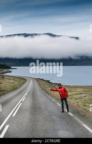 Männlicher Tourist in Oberbekleidung, der am Straßenrand steht und auf dem Anhalter steht Hintergrund der majestätischen Landschaft von Bergen und See Stockfoto