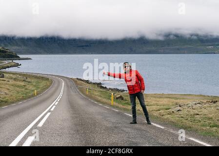 Männlicher Tourist in Oberbekleidung, der am Straßenrand steht und auf dem Anhalter steht Hintergrund der majestätischen Landschaft von Bergen und See Stockfoto