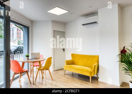Moderne einfache Innenarchitektur von hellen gemütlichen geräumigen Büro mit Bequeme orangefarbene Stühle an einem runden Holztisch und einem gelben Sofa Gegen weiße Wände und große Fenster Stockfoto
