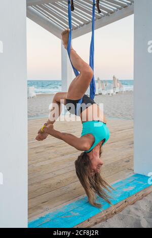 Sportliche Power-Frau, die kopfüber auf der Hängematte hängt, um sich am Bein in einem hölzernen Stand am Strand Yoga aus der Luft zu halten Stockfoto