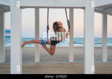 Fröhliche Frau, die sich das Bein auf der blauen Hängematte für Luft-Yoga auf der hölzernen Bühne streckt Stockfoto