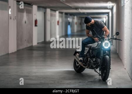 Ganzkörper bärtigen Kerl in Freizeitkleidung sitzt auf Motorrad Und Blick weg in Korridor der modernen Garage Stockfoto