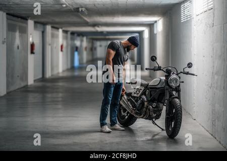 Ganzkörper bärtigen Kerl in lässigen Kleidung in der Nähe auf stehen Motorrad im Korridor der modernen Garage Stockfoto